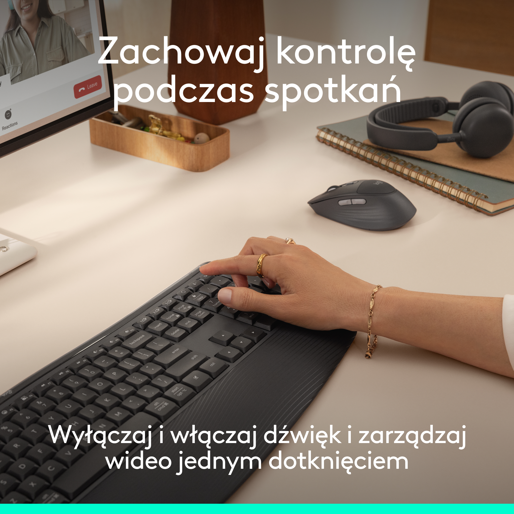 Dłoń pisze na klawiaturze, obok mysz, słuchawki i laptop z wideokonferencją.