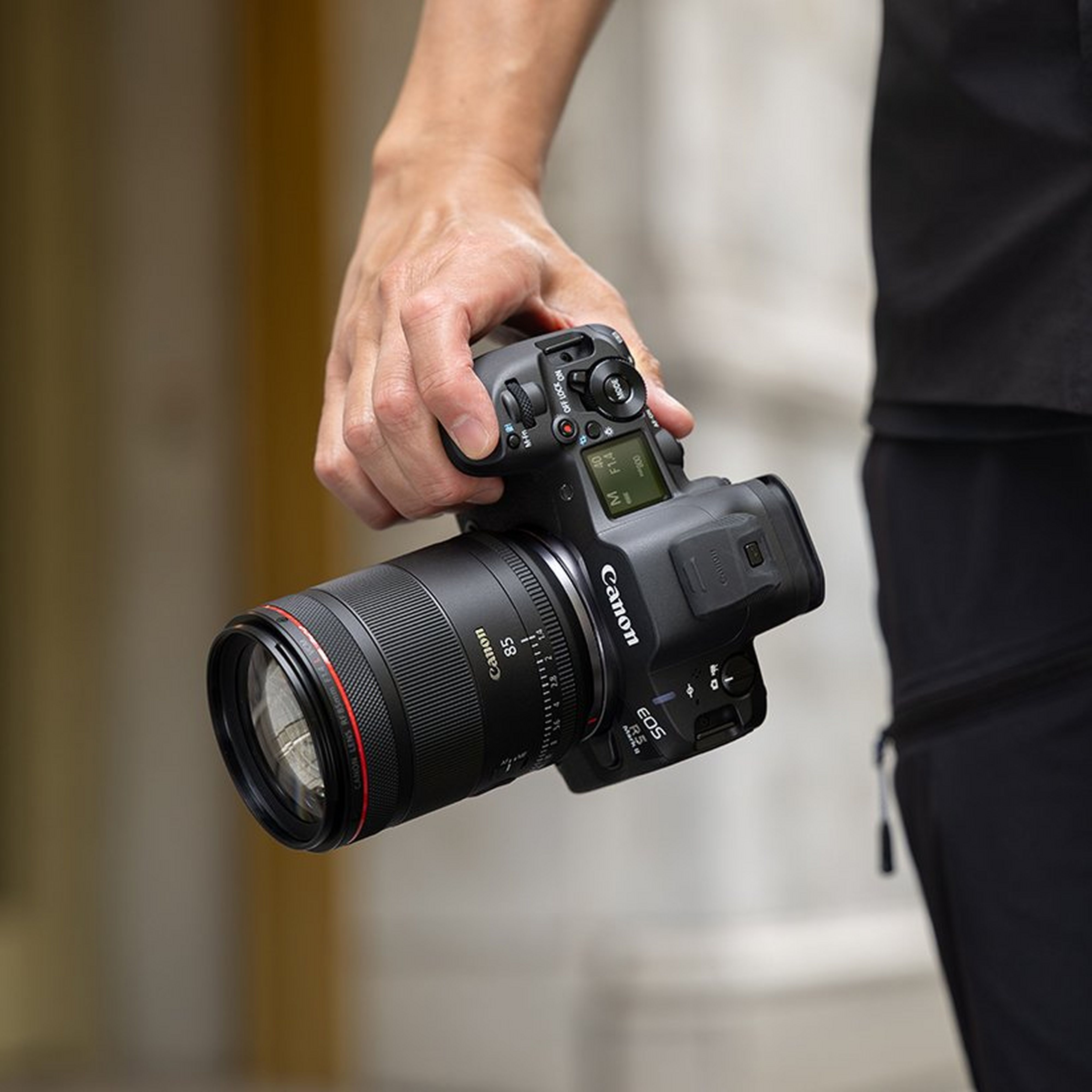 Una persona tiene una fotocamera Canon EOS R5 nera con obiettivo da 85mm. La fotocamera mostra impostazioni.