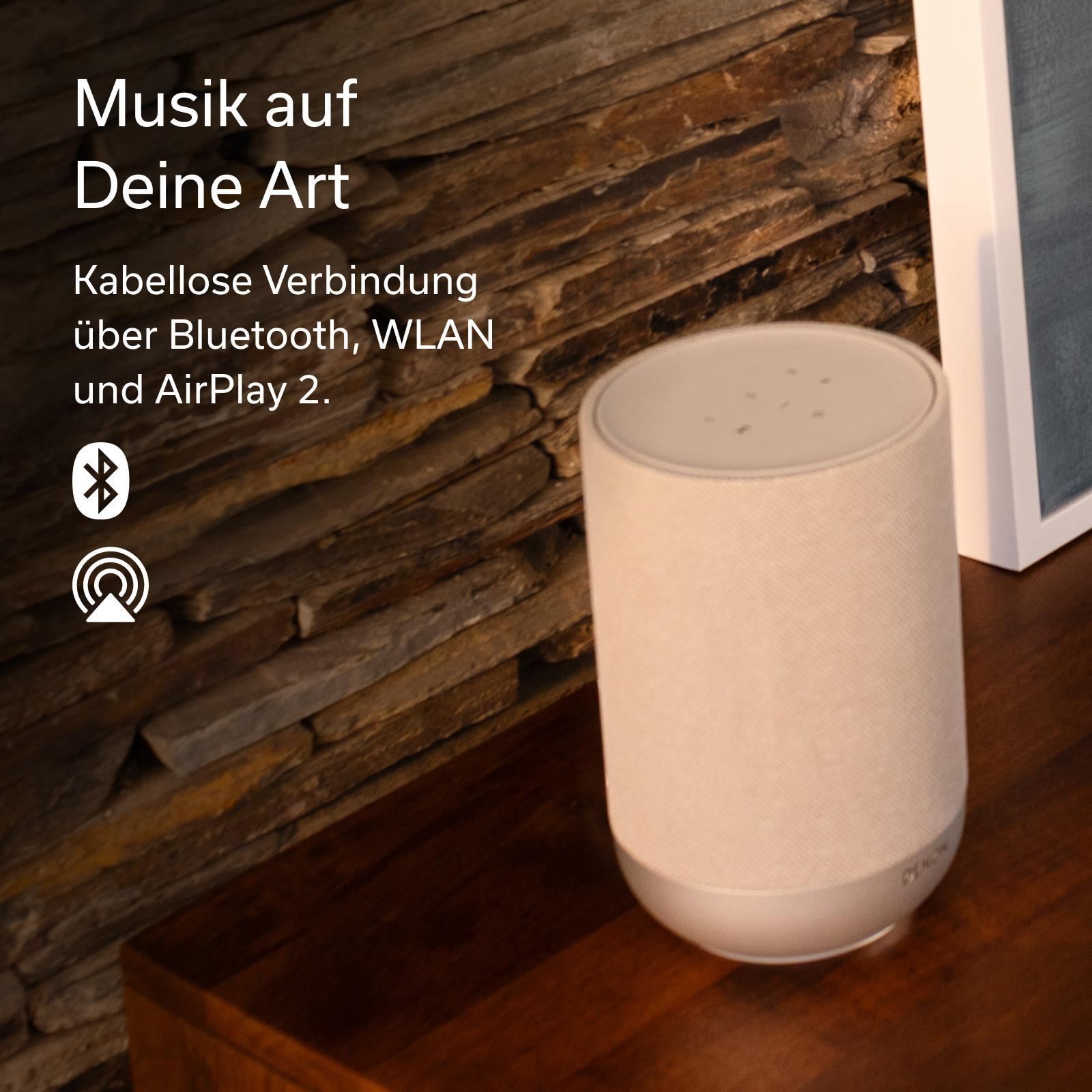 Ein weißer Lautsprecher steht auf einem Holztisch vor einer Steinwand.