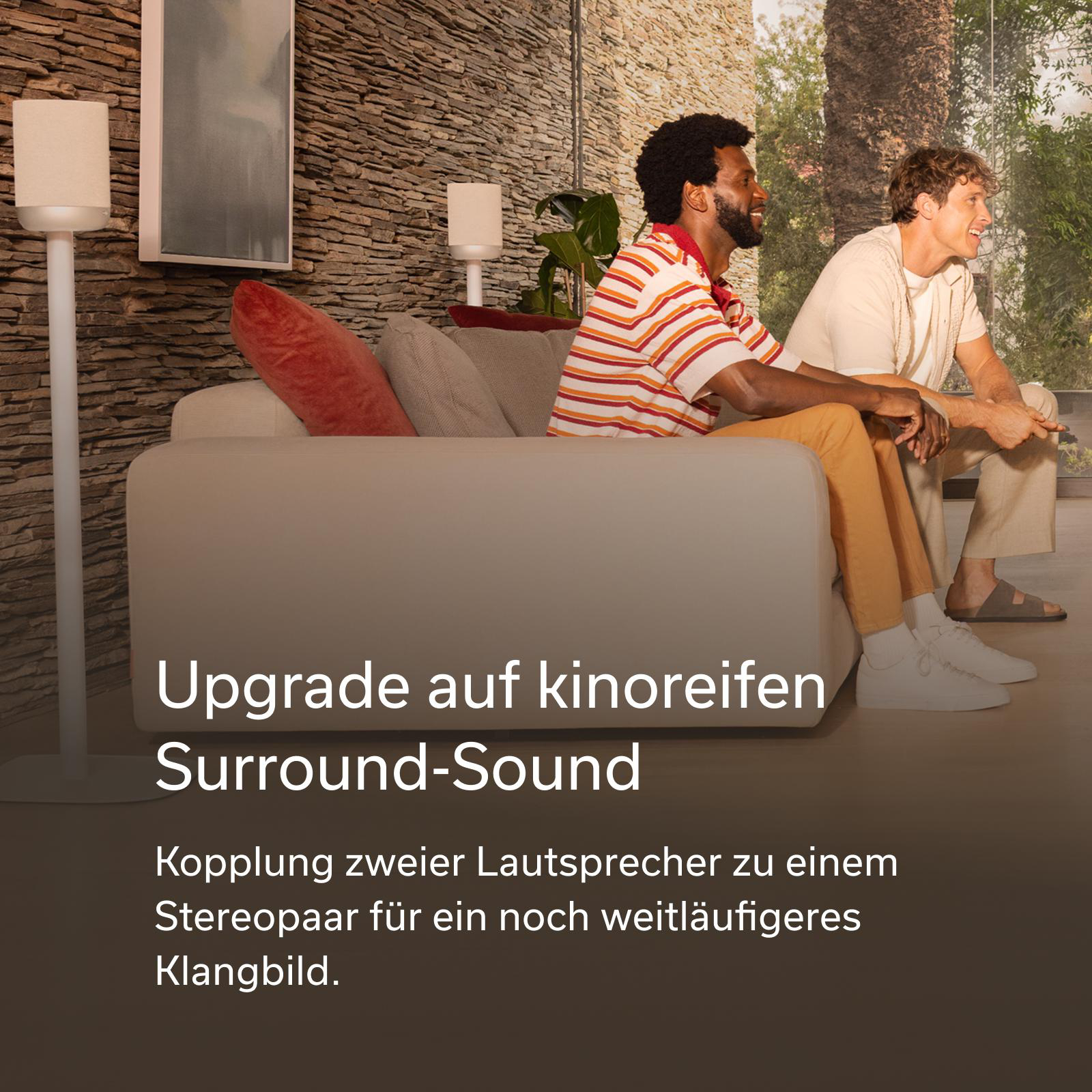Zwei Männer sitzen auf einem Sofa vor einer Steinwand. Sie schauen nach rechts.