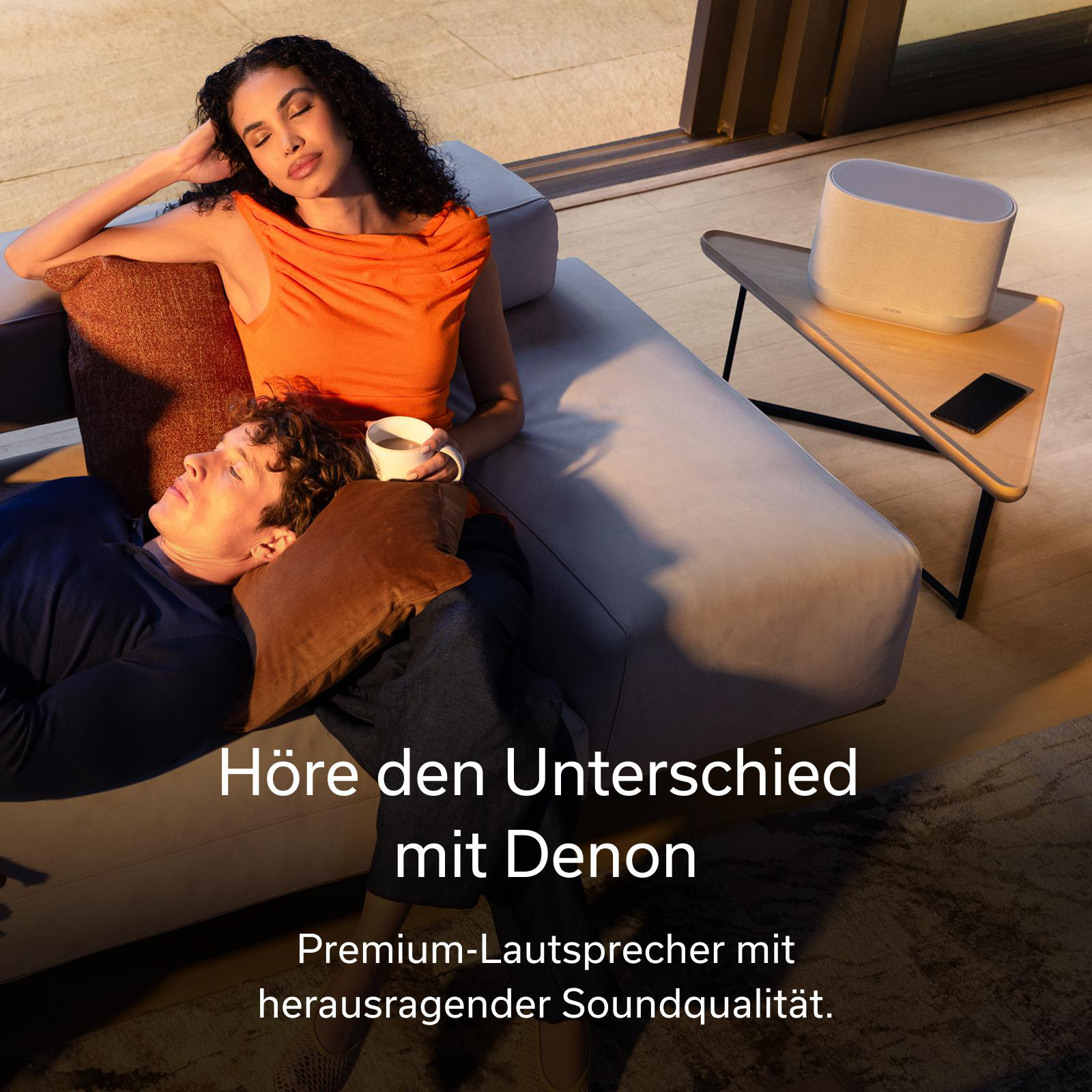 Frau mit orangenem Oberteil und Mann auf Sofa, Lautsprecher auf Tisch.