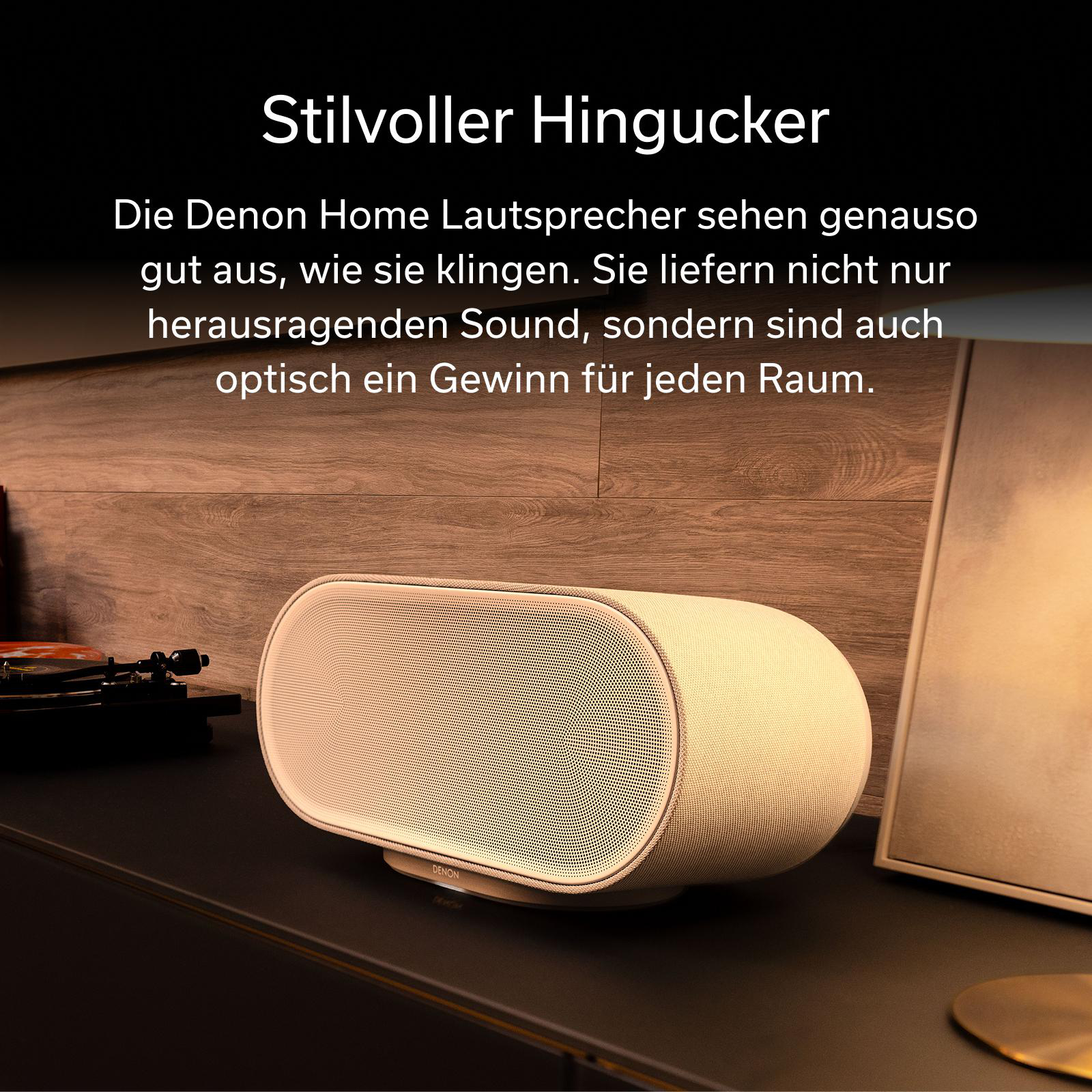 Ein beiger Denon-Lautsprecher steht auf dunkler Oberfläche vor Holz Hintergrund. Links ist ein Plattenspieler.