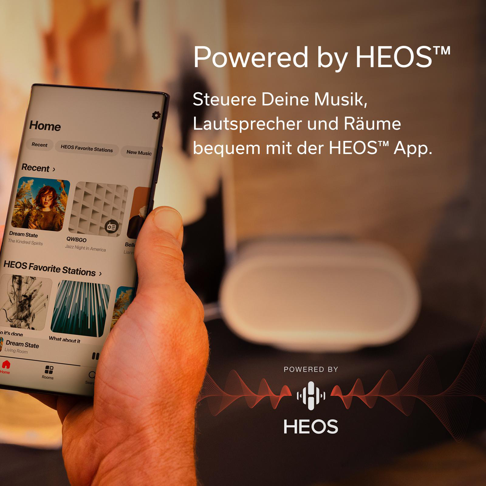 Ein Smartphone zeigt die HEOS App mit Musik und Räumen. Rechts steht "Powered by HEOS".
