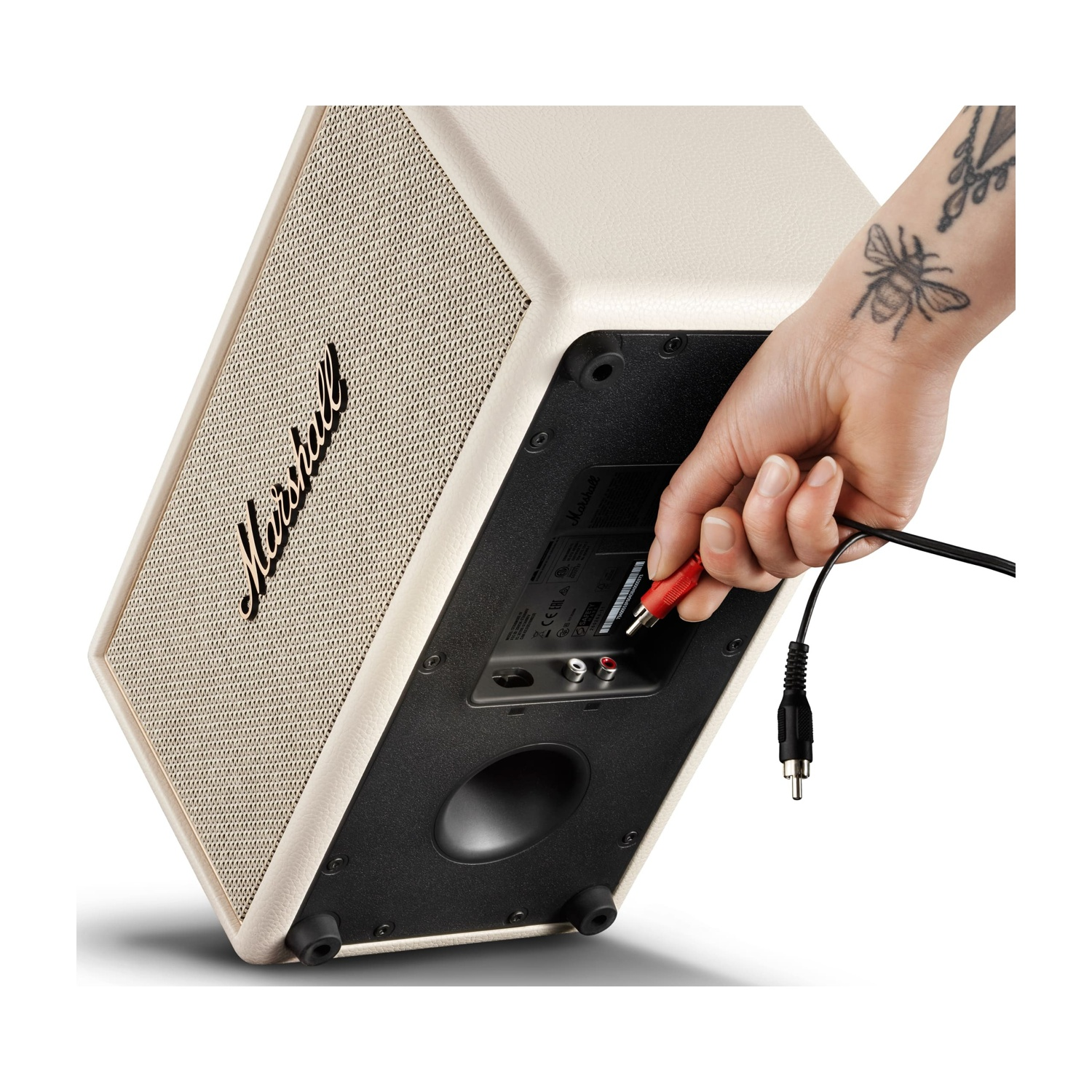 Una mano con tatuaggio di ape inserisce un cavo audio rosso in un altoparlante color crema.