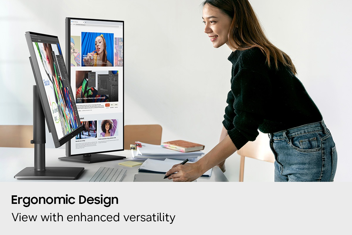 Una donna in jeans e maglione nero lavora a una scrivania con doppio monitor che mostra siti web.