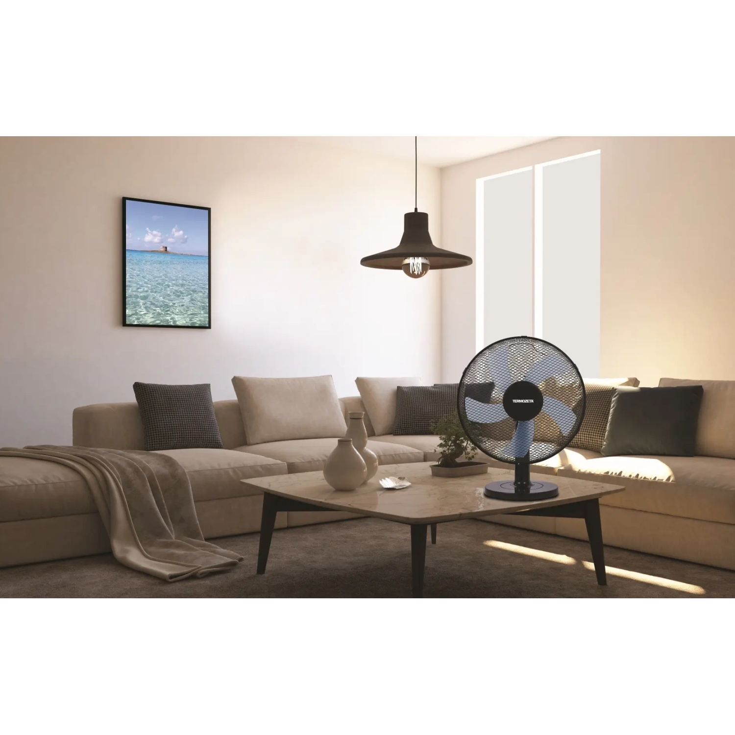 A fan sits on a coffee table in front of a sofa. A picture of a tower in the sea hangs on the wall.