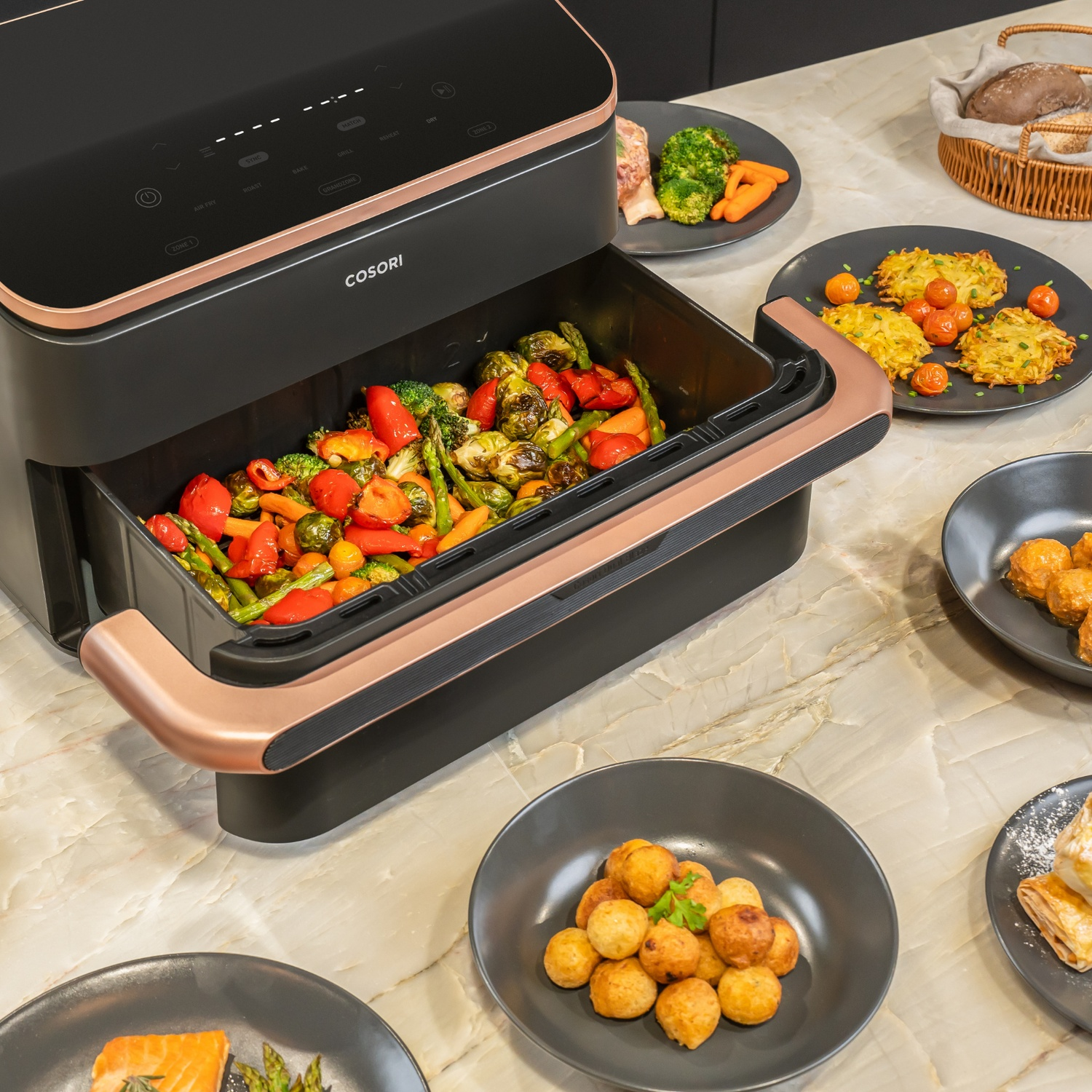 Friteuse à air noire avec accents de cuivre, remplie de légumes rôtis. Plusieurs assiettes de nourriture autour.