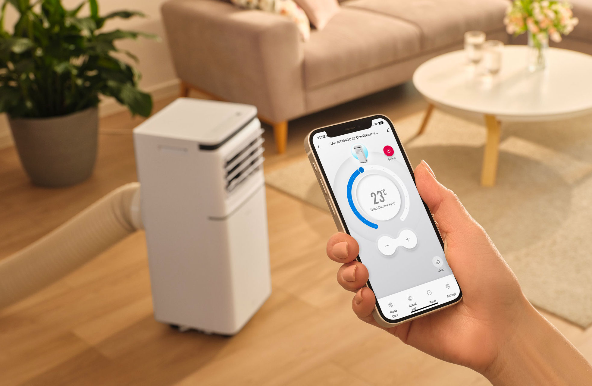 Biały przenośny klimatyzator z wężem stoi na drewnianej podłodze, obok telefonu.
