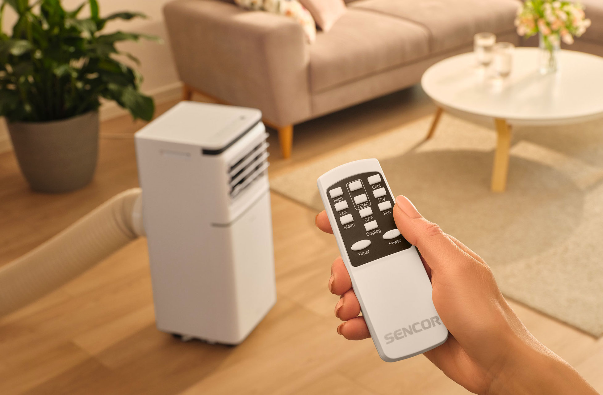 Dłoń trzyma pilot Sencor sterujący przenośnym klimatyzatorem w salonie.