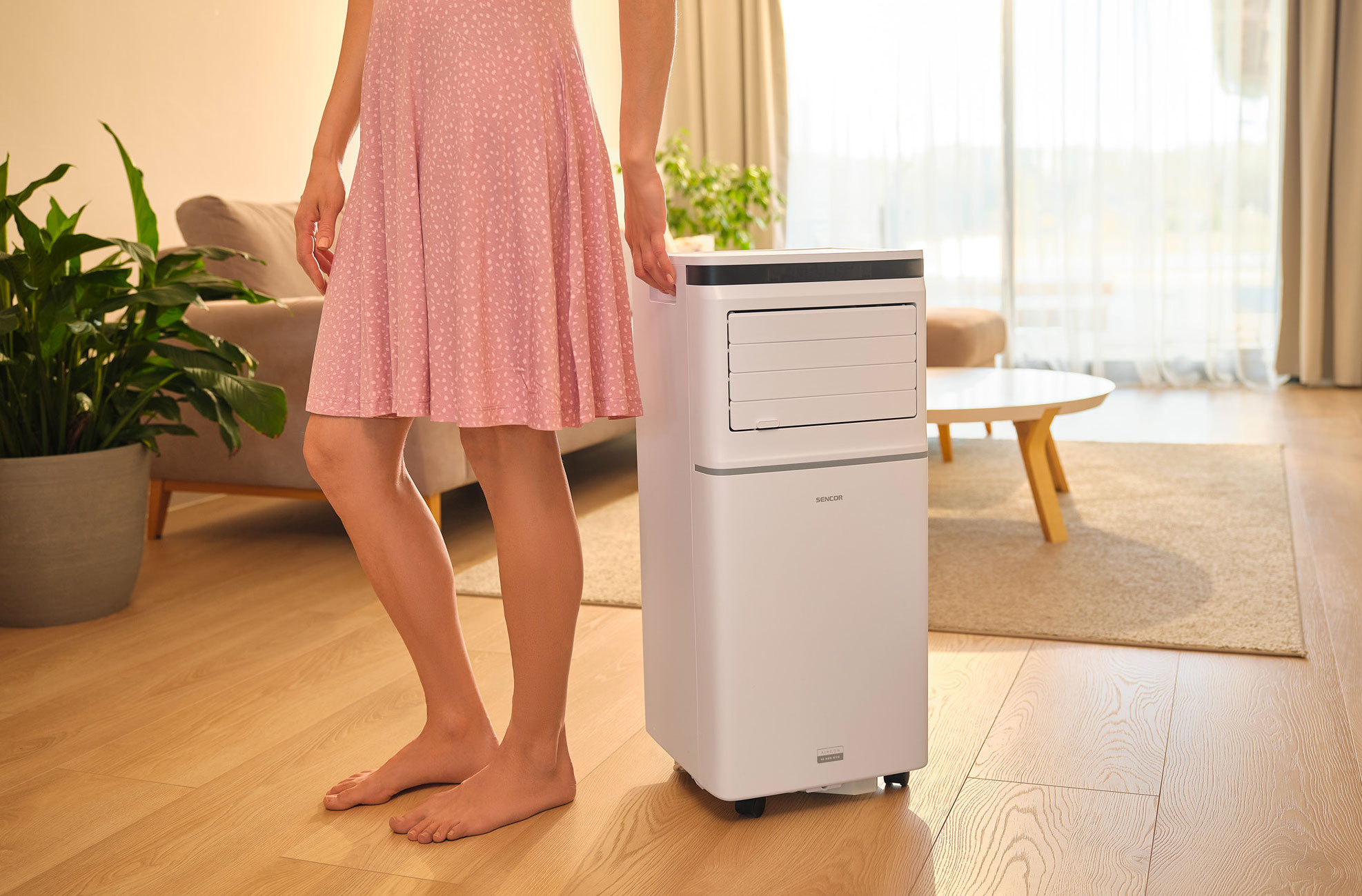 Kobieta w różowej sukience stoi obok białej klimatyzacji w salonie.