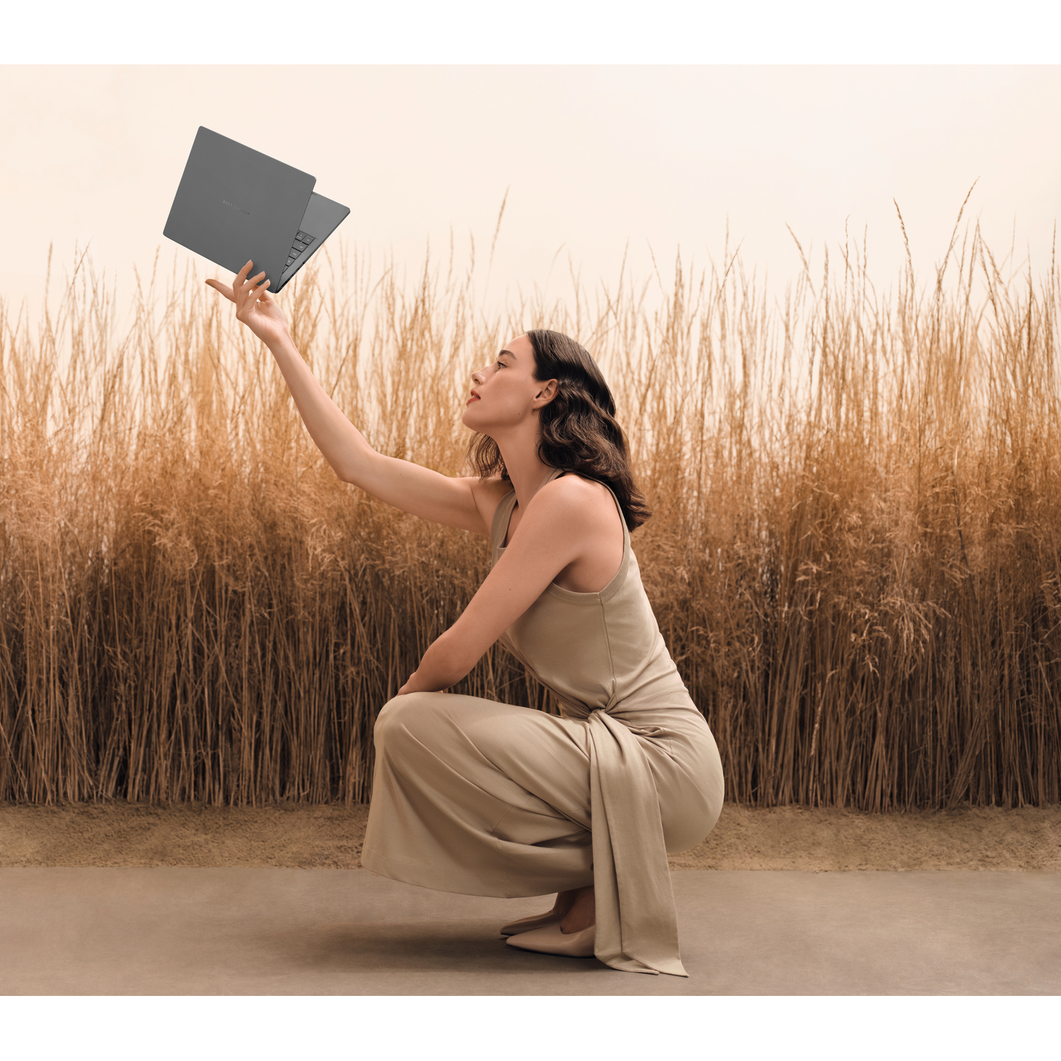 Donna in pantaloni e top beige tiene un laptop grigio sopra steli d'erba dorata.