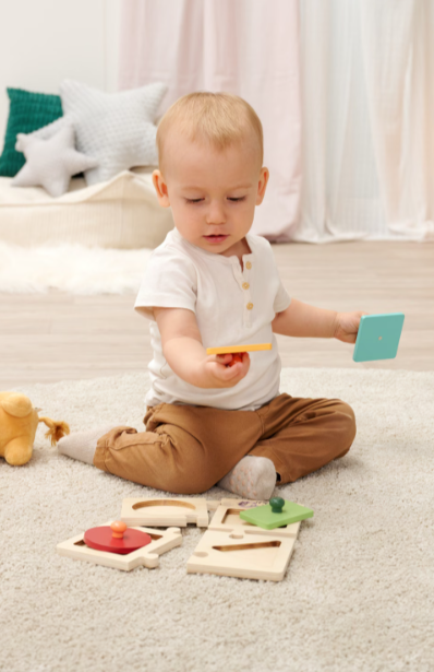 Ein Baby spielt mit Holzpuzzles auf einem Teppich. Im Hintergrund sind Kissen.