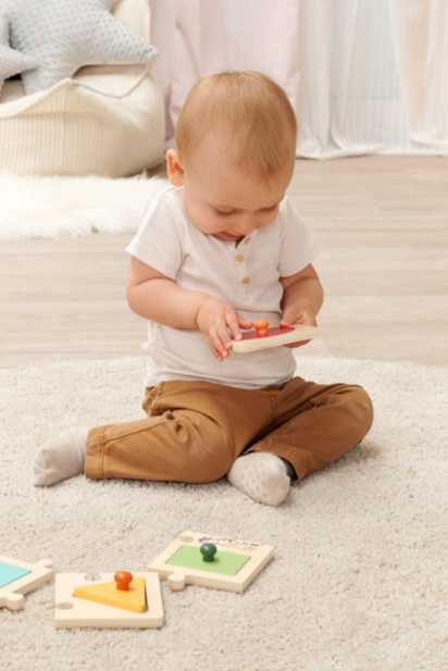 Ein Baby in brauner Hose und weißem Hemd spielt mit Holzpuzzles auf einem flauschigen Teppich.