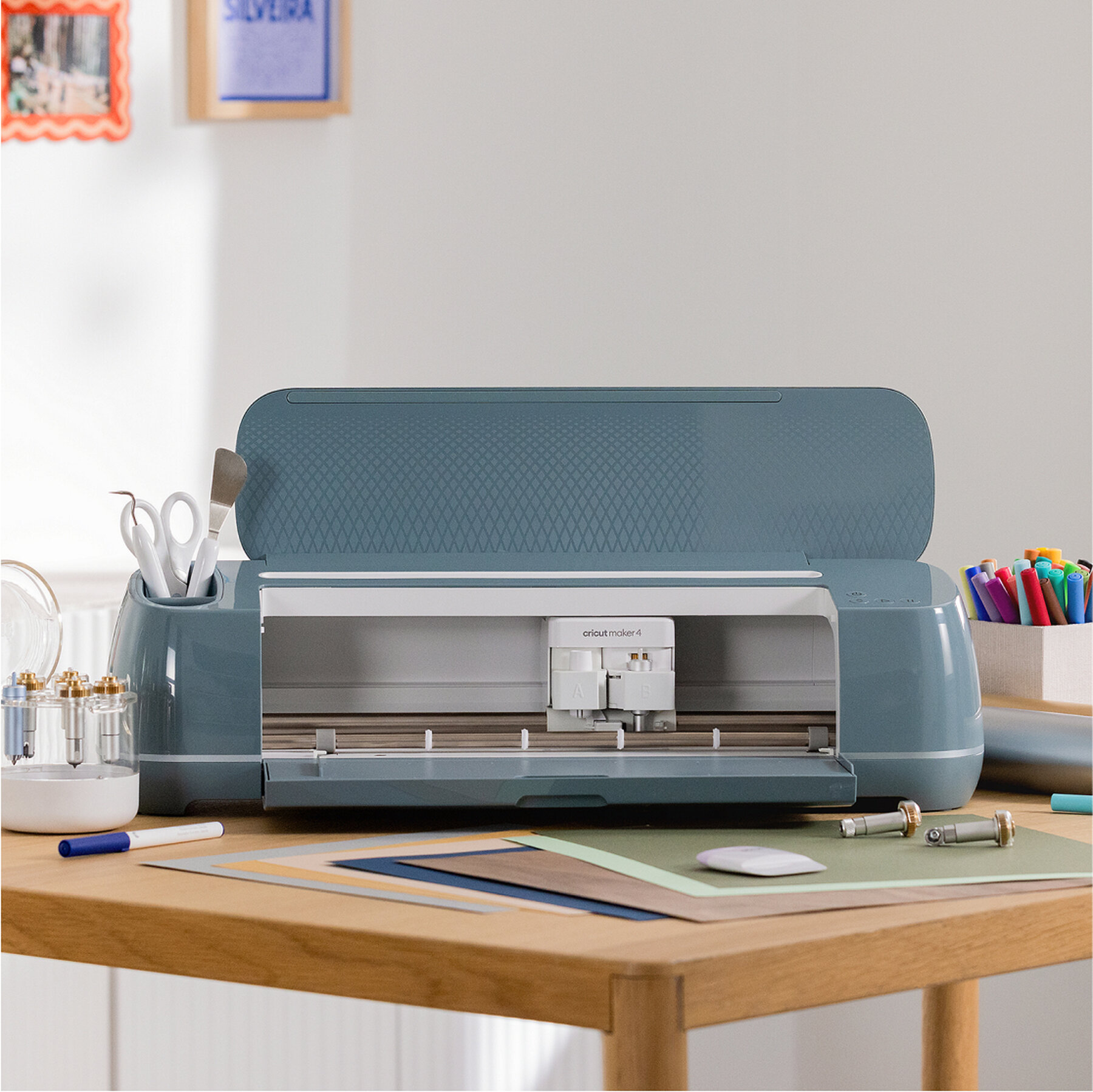 Ahşap bir masa üzerinde aletler ve renkli kalemler bulunan mavi bir Cricut Maker 4.