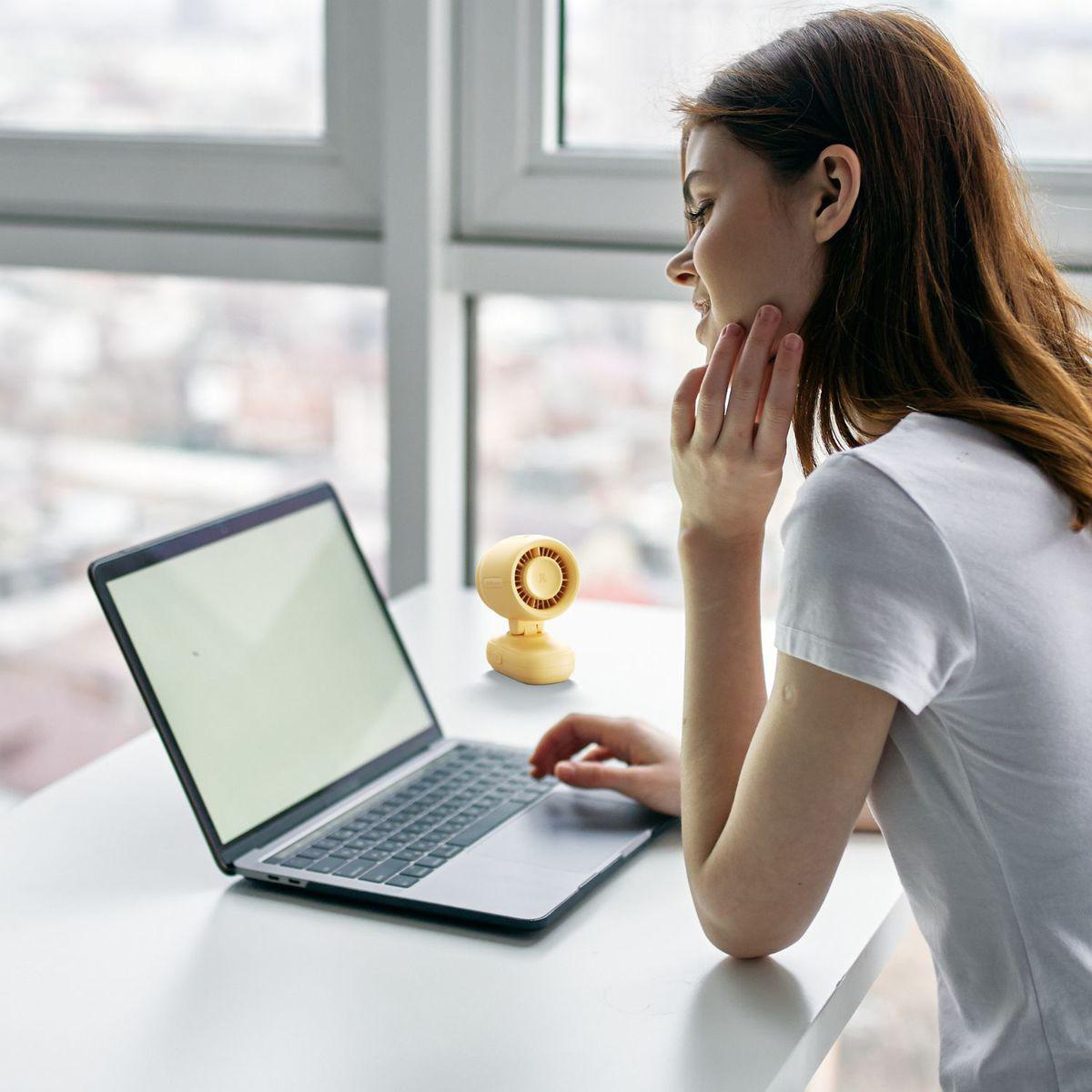 Eine Frau sitzt am Laptop, ein kleiner gelber Ventilator steht daneben.