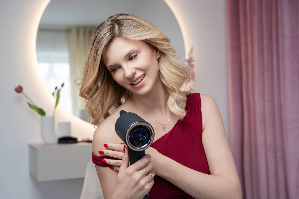 Kobieta z blond włosami używa ciemnej suszarki do włosów. Nosi czerwoną sukienkę.
