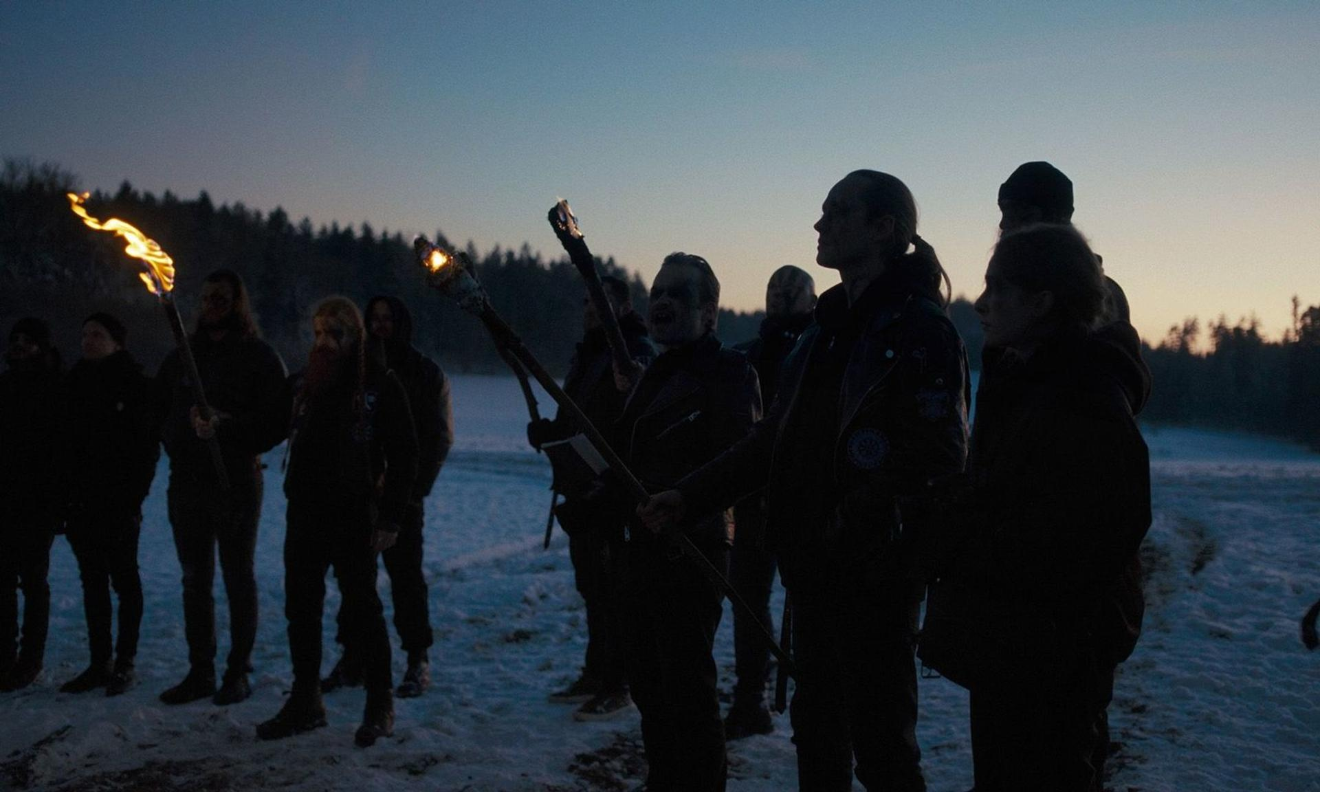 Gruppe von Menschen mit Fackeln auf einem verschneiten Feld in der Dämmerung, Wald im Hintergrund.
