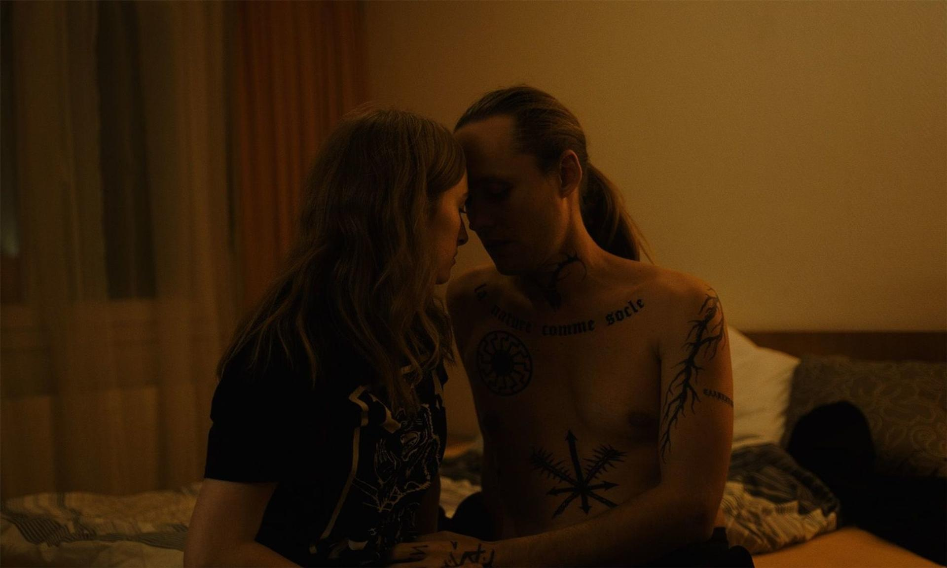 Ein Mann mit Tattoos und eine Frau sitzen eng beieinander auf einem Bett.