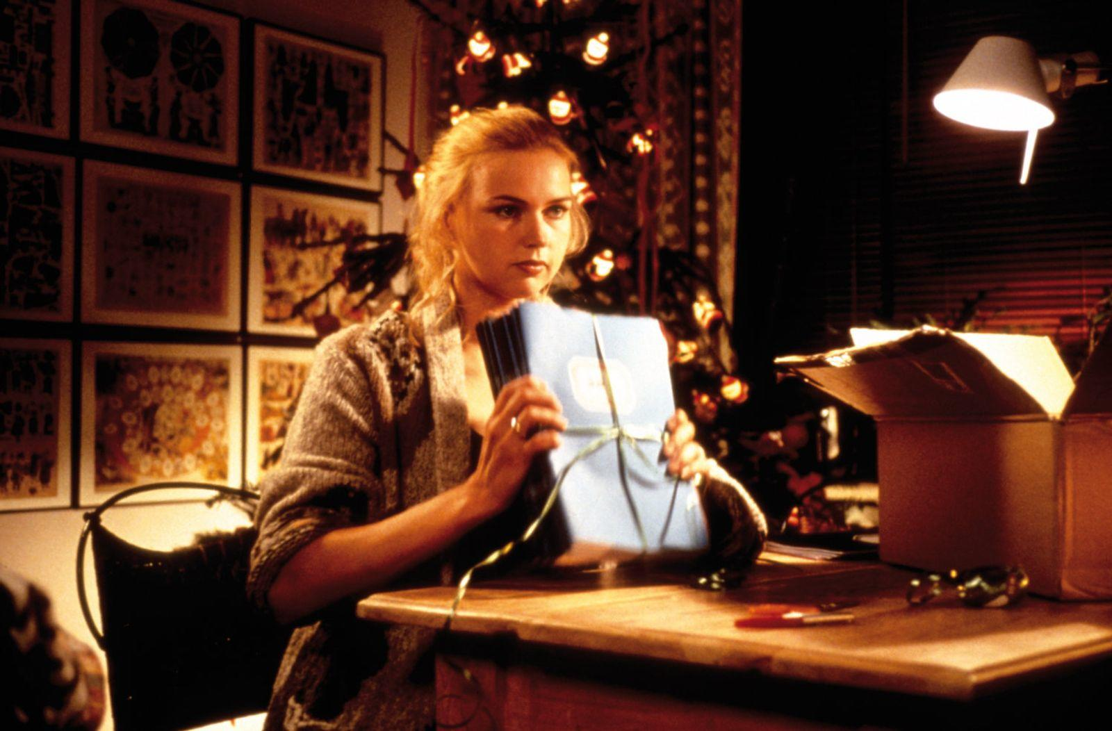 Eine blonde Frau in einem grauen Cardigan hält ein hellblaues Geschenkpaket, das mit einem grünen Band gebunden ist, an einem Holzschreibtisch sitzend.