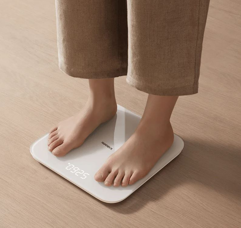 Osoba stoi na białej wadze cyfrowej pokazującej 52,60 kg. Stopy są nagie.