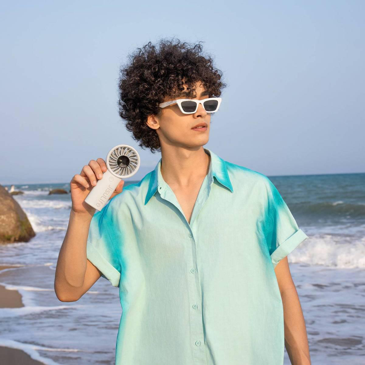 Mann mit lockigem Haar und Sonnenbrille hält Lüfter am Strand mit Meer im Hintergrund.
