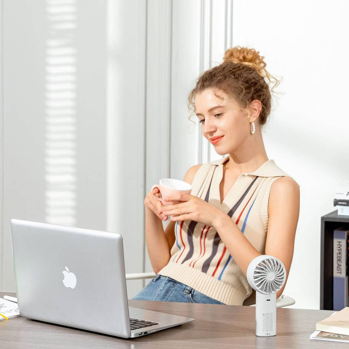 Eine Frau sitzt an einem Schreibtisch mit einem Laptop und trinkt aus einer Tasse, ein kleiner Ventilator steht daneben.