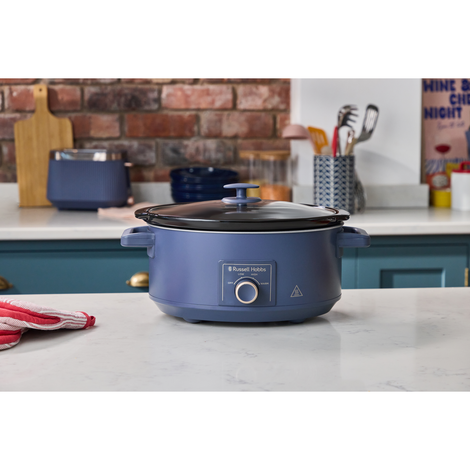Een blauwe slowcooker staat op een marmeren aanrecht met ernaast een rood-witte ovenwant.