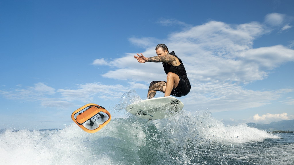 Ein Mann auf einem Surfbrett springt über eine Welle, neben ihm fliegt eine orangefarbene Drohne.