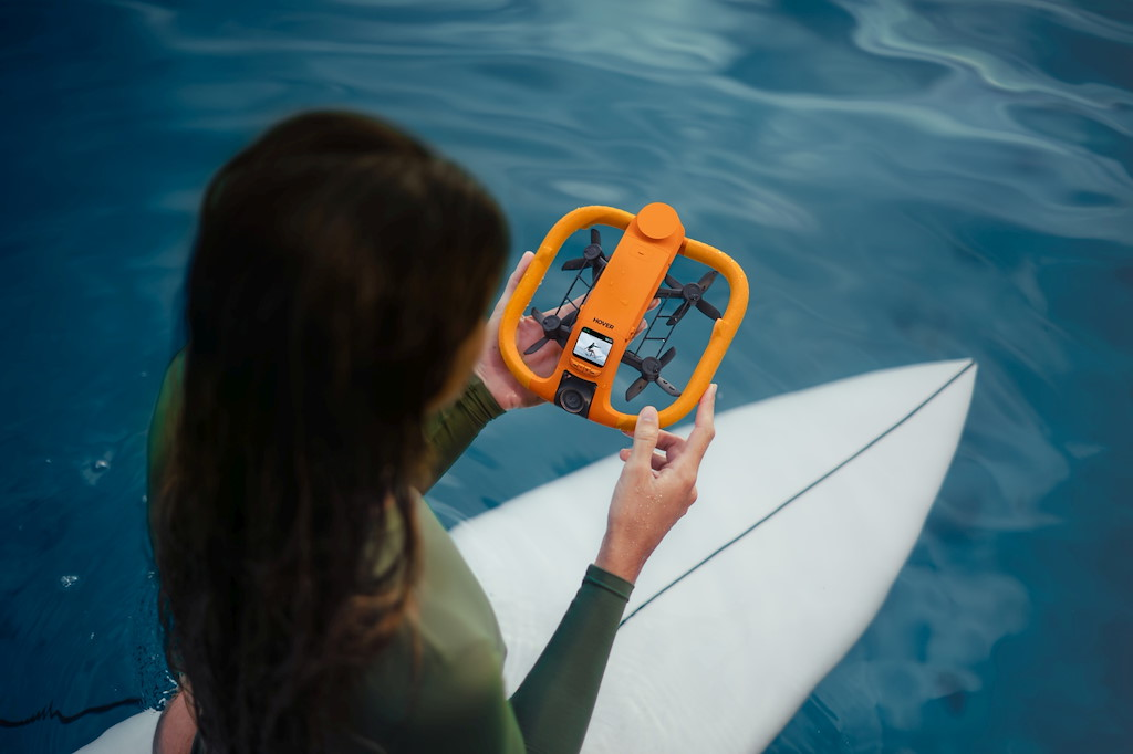 Eine Person hält eine orangefarbene Drohne über einem Surfbrett im Wasser.