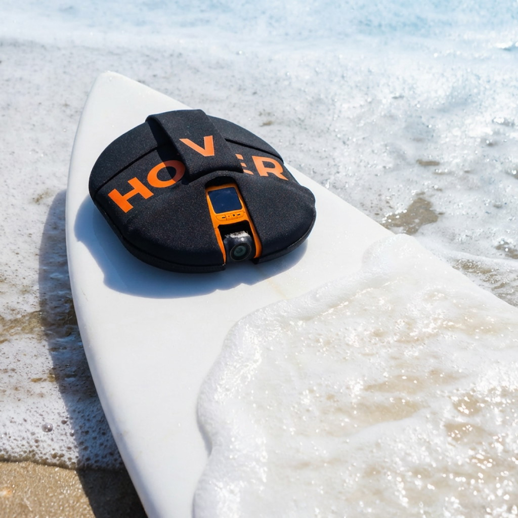 Ein schwarzes Hoverboard mit orangefarbener Schrift liegt auf einem weißen Surfbrett am Strand.