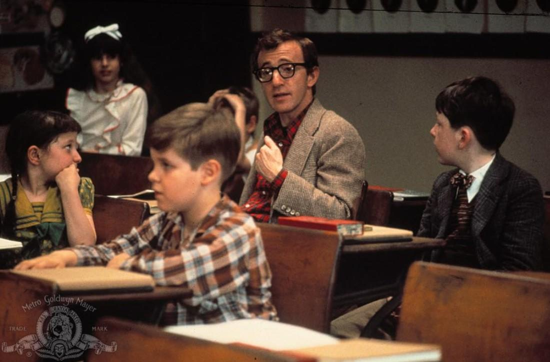 Ein Mann mit Brille in einem karierten Hemd und Sakko spricht zu Kindern in einem Klassenzimmer.
