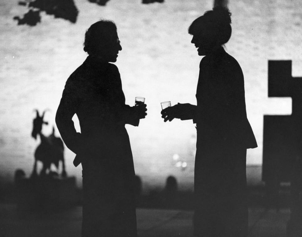 Silhouetten zweier Personen mit Gläsern, die sich unterhalten. Im Hintergrund eine Statue.