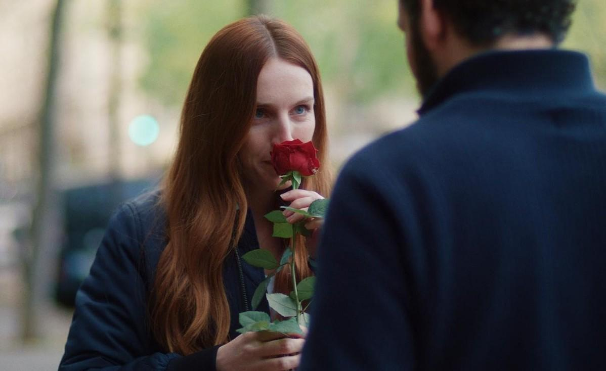 Eine Frau mit roten Haaren riecht an einer roten Rose, ein Mann steht ihr gegenüber.