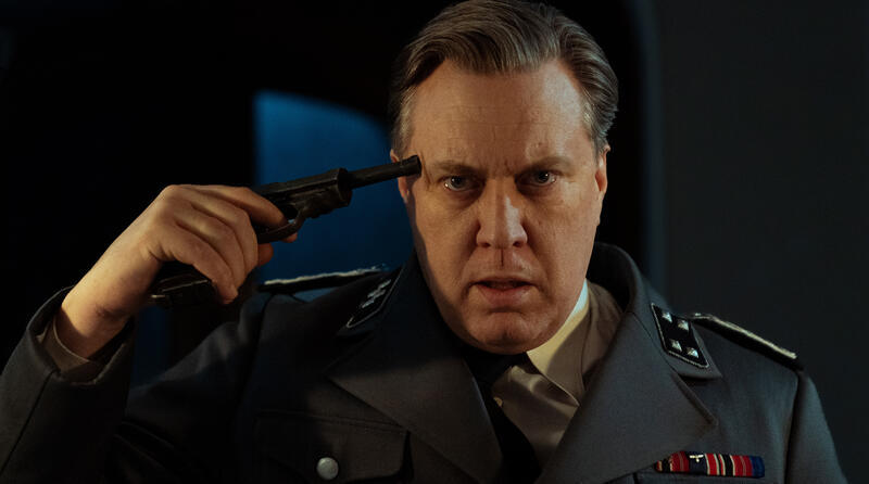 Mann in Uniform hält Pistole an seine Schläfe, dunkler Hintergrund.
