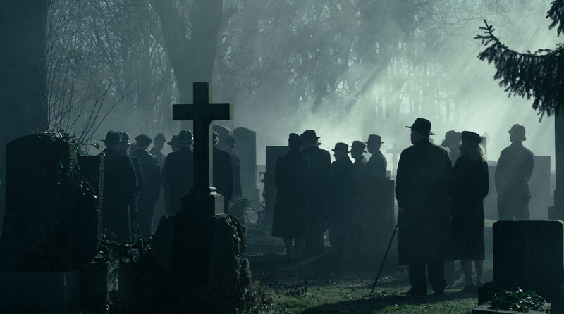 Eine Gruppe von Menschen in Hüten und Mänteln steht auf einem nebligen Friedhof.