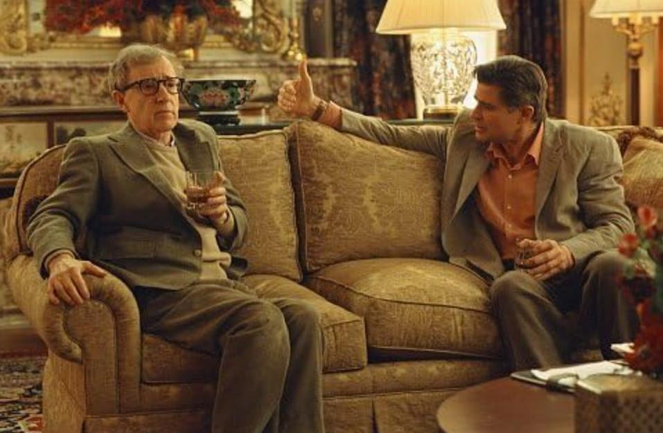 Zwei Männer sitzen auf einem Sofa. Einer hält ein Glas, der andere macht eine Daumen-hoch-Geste.
