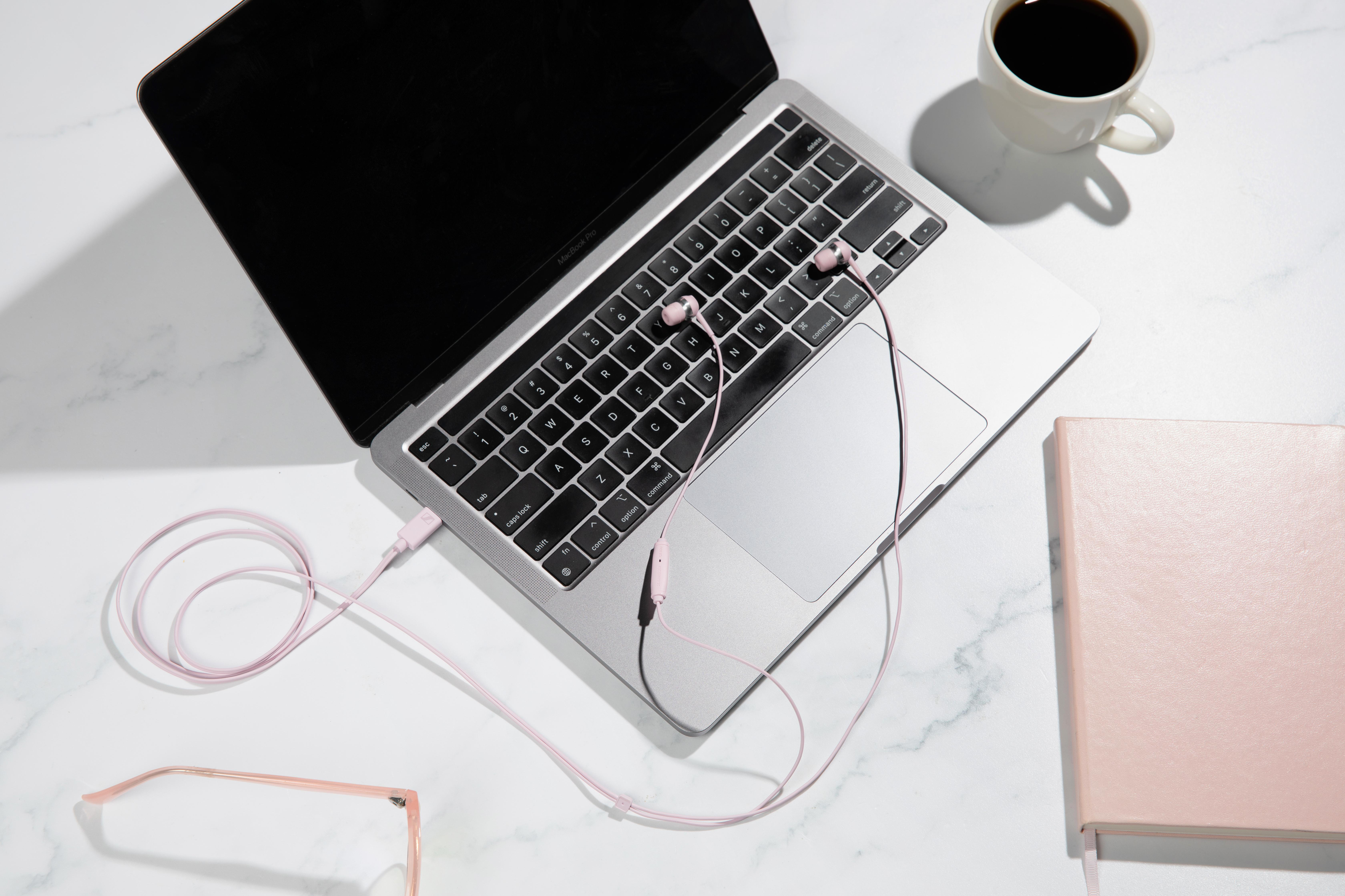 Ein offener Laptop mit rosa Kopfhörern, einer Kaffeetasse und einem Notizbuch.