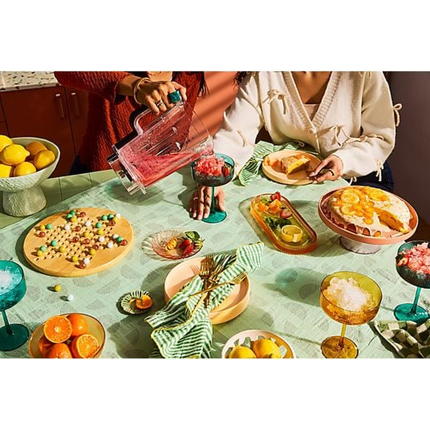 A table with fruit, cake, candy, and drinks, people are preparing something.
