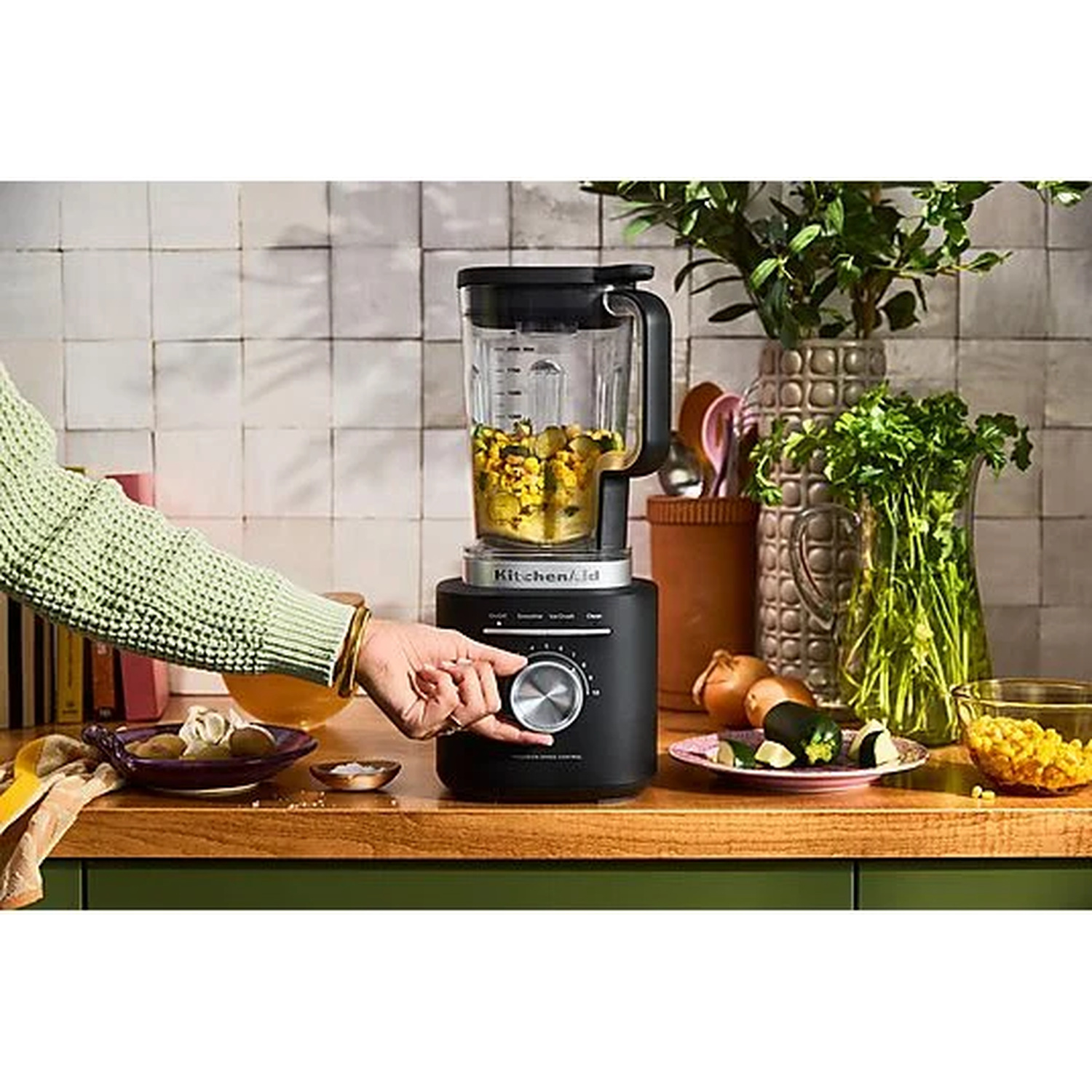 A hand adjusts a black KitchenAid blender filled with corn and chickpeas. Ingredients on counter.