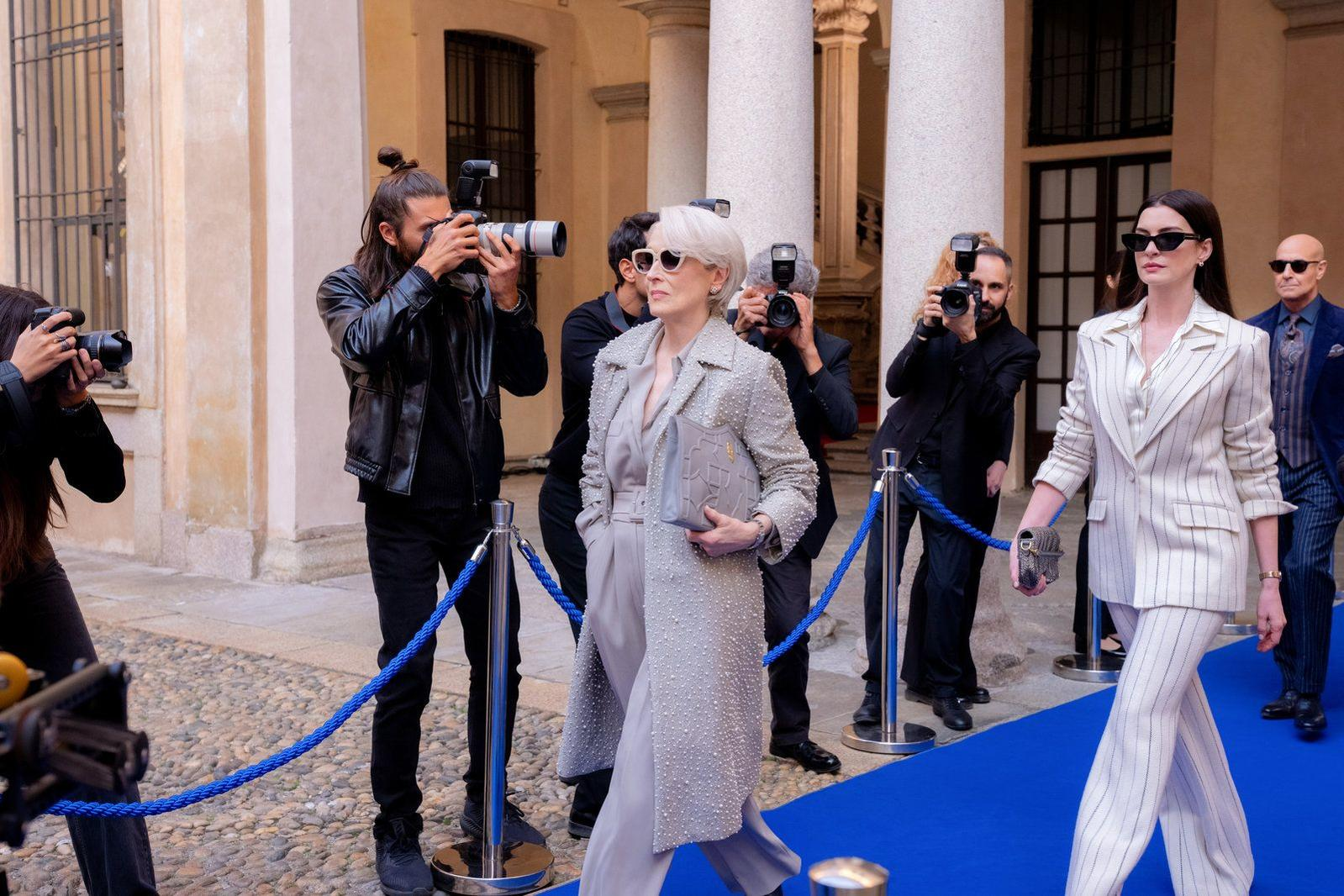 Paparatzi fotografieren zwei Frauen auf einem blauen Teppich vor einem Steingebäude.