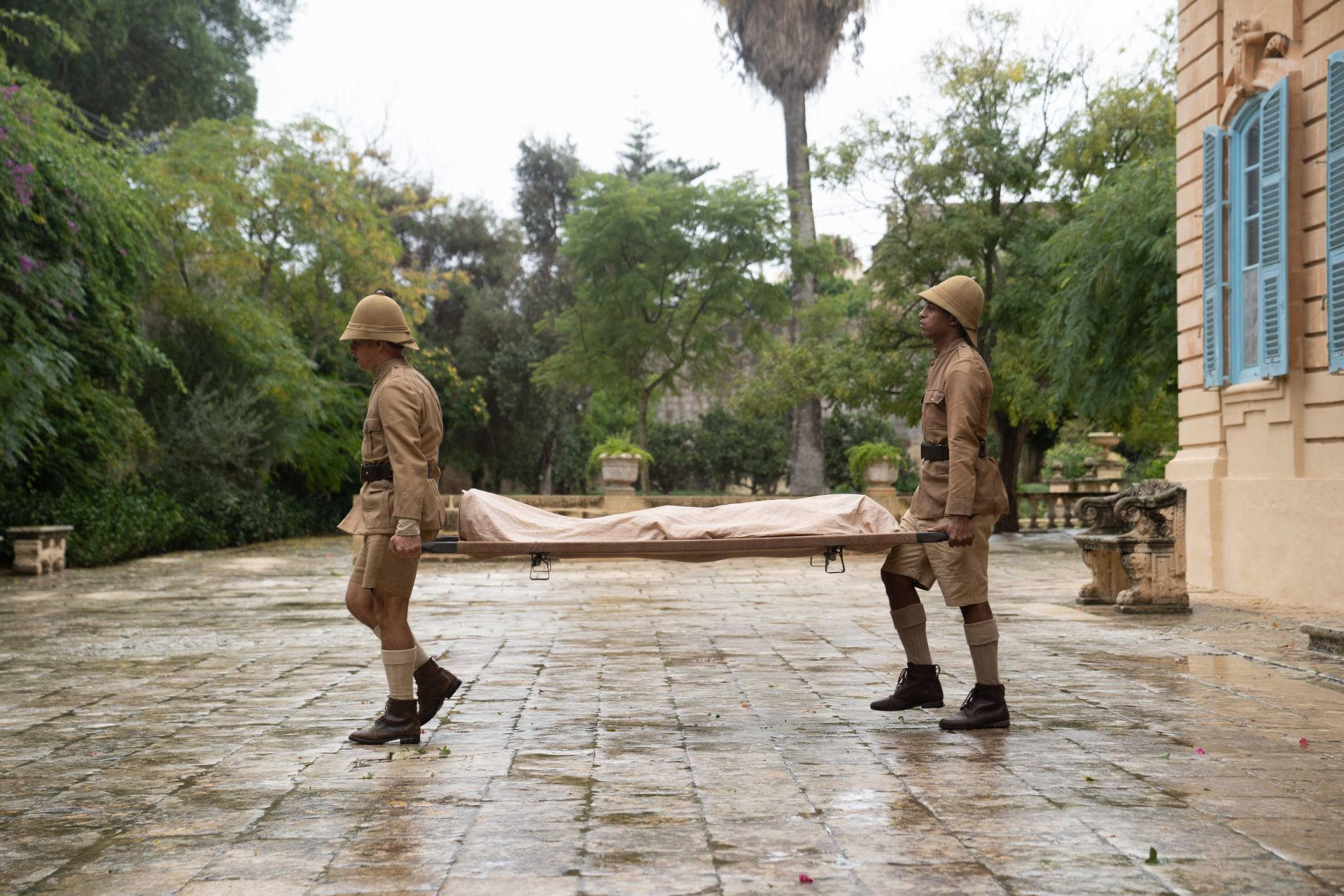 Zwei Soldaten in Khaki-Uniformen tragen eine Trage mit einer Leiche durch einen nassen Hof.