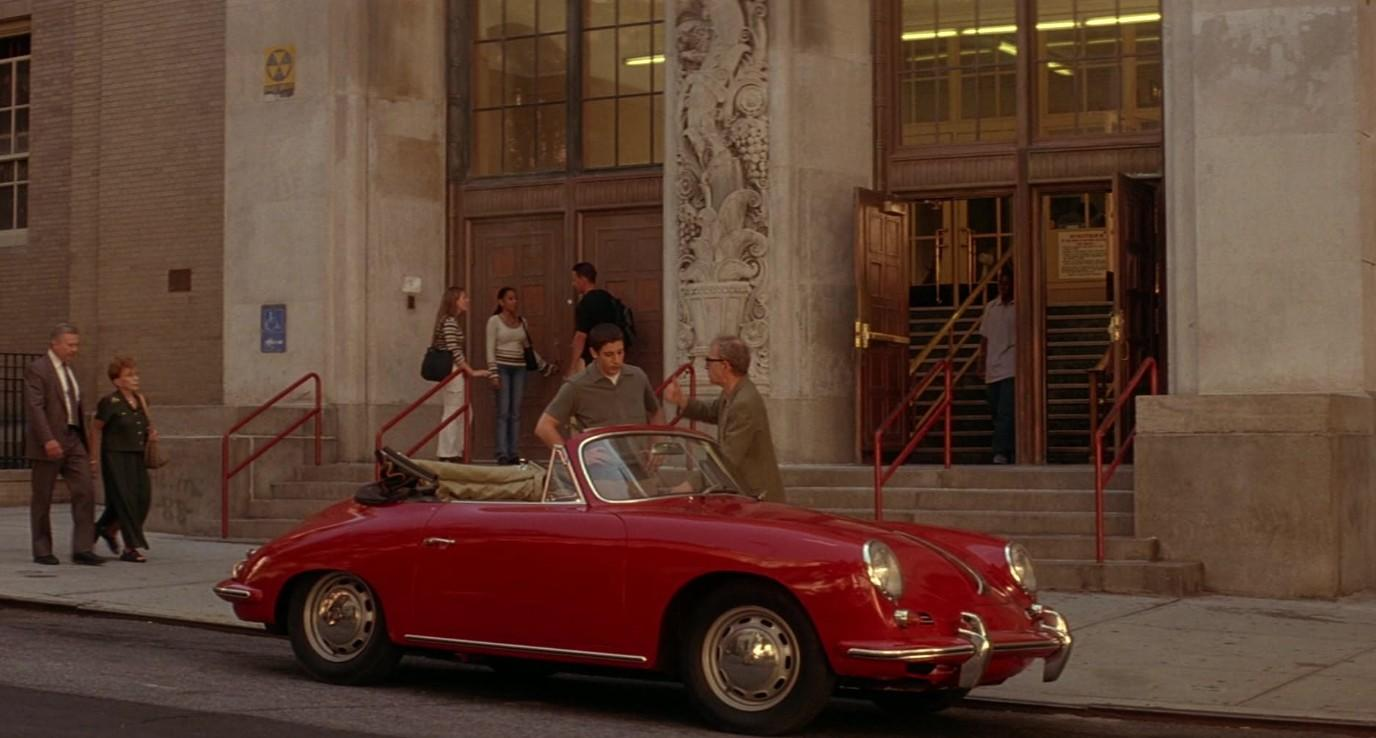 Ein rotes Cabrio parkt auf einer Straße vor einem Gebäude mit Treppen und eintretenden Menschen.
