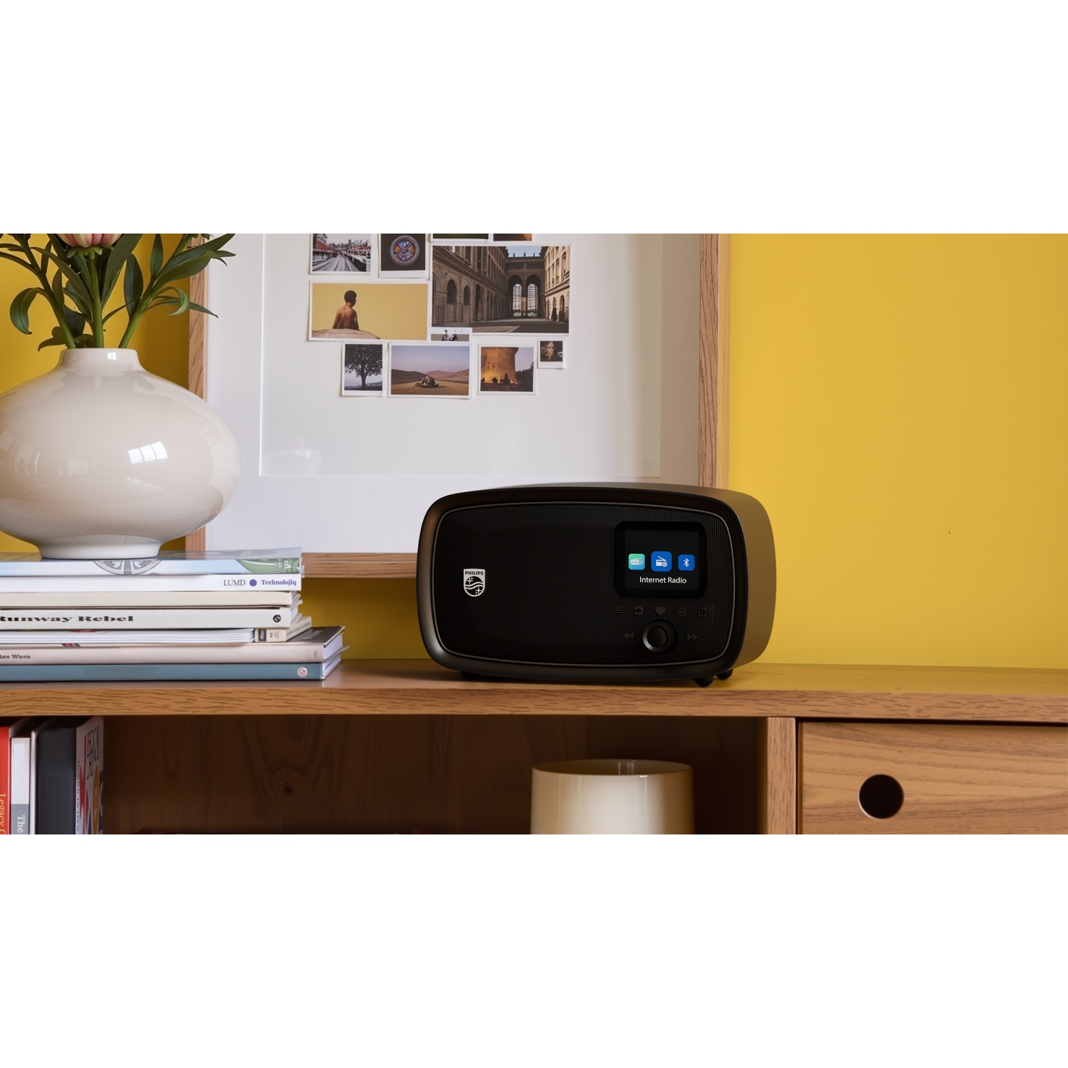 Radio internet nera su mensola in legno con libri e vaso di fiori.
