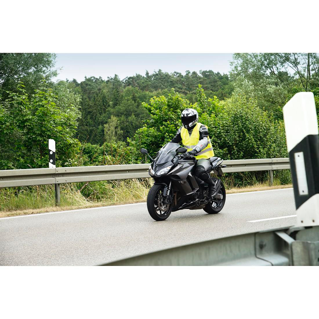 Ein Motorradfahrer in Warnweste fährt auf einer Straße, umgeben von Bäumen.
