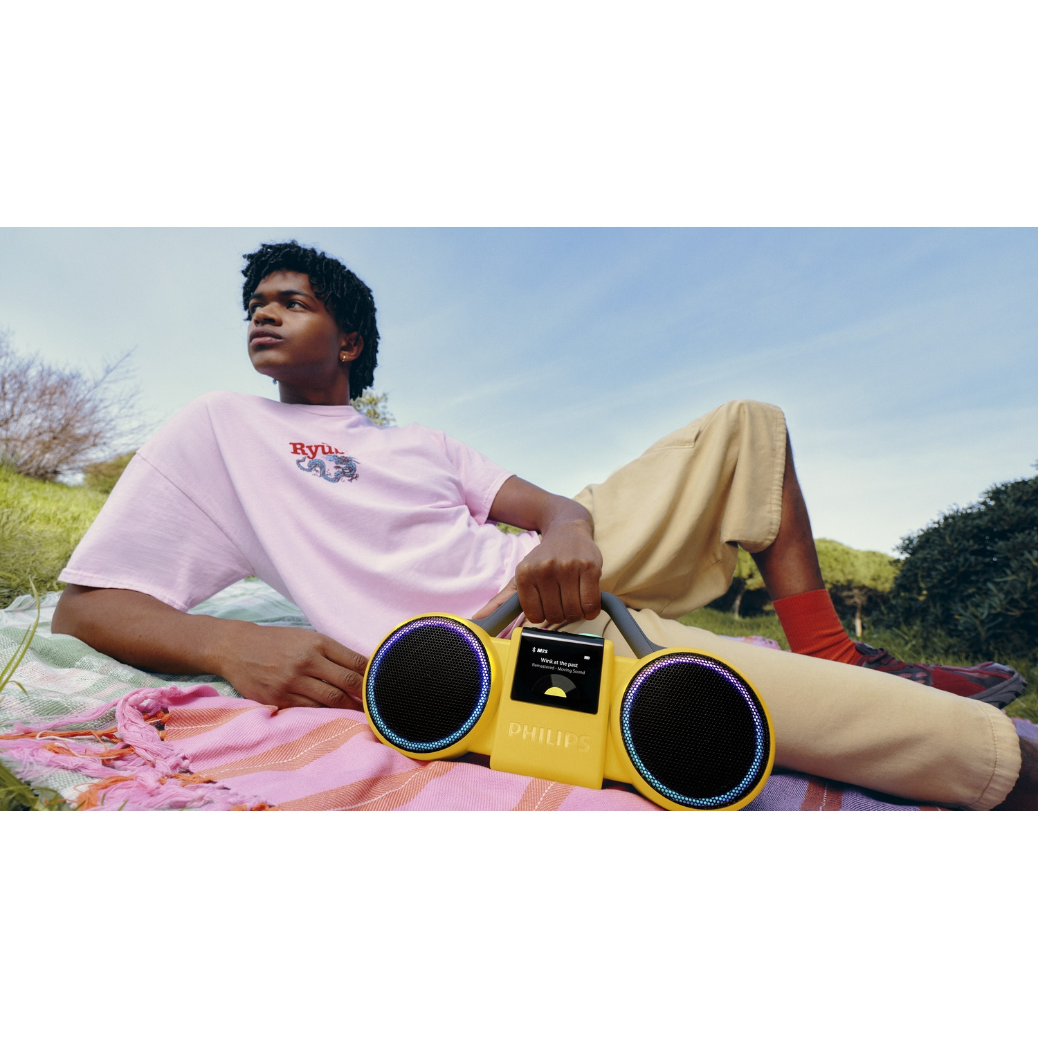 Un uomo giace su una coperta all'aperto, tenendo uno speaker Philips giallo con luci.