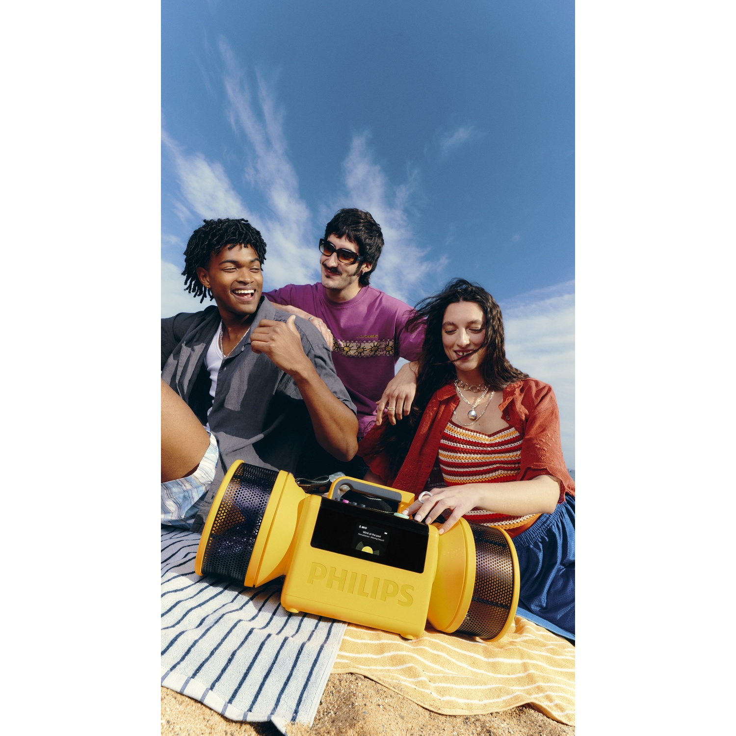 Tre amici seduti sulla spiaggia con uno speaker Philips giallo.