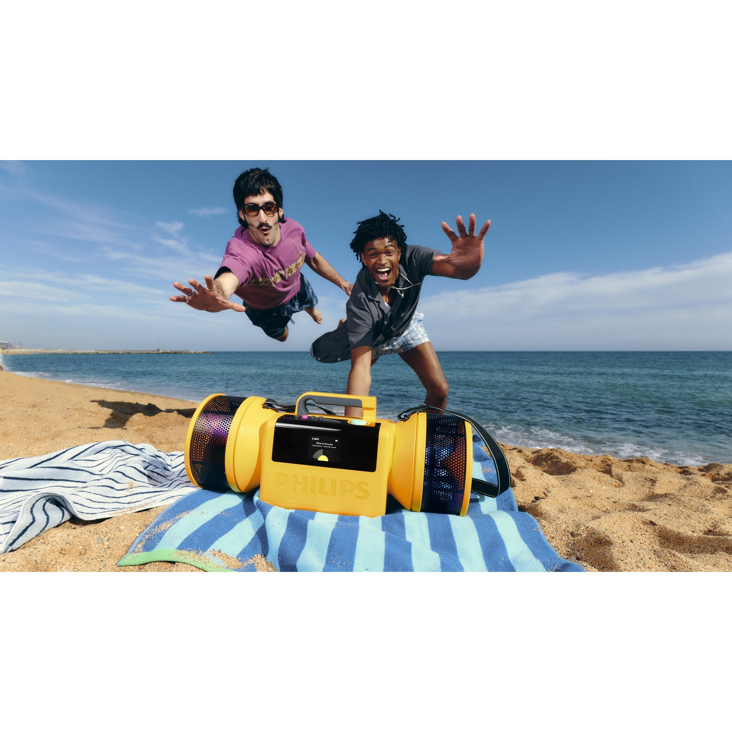 Due uomini saltano su una spiaggia davanti a uno speaker Philips giallo e al mare.