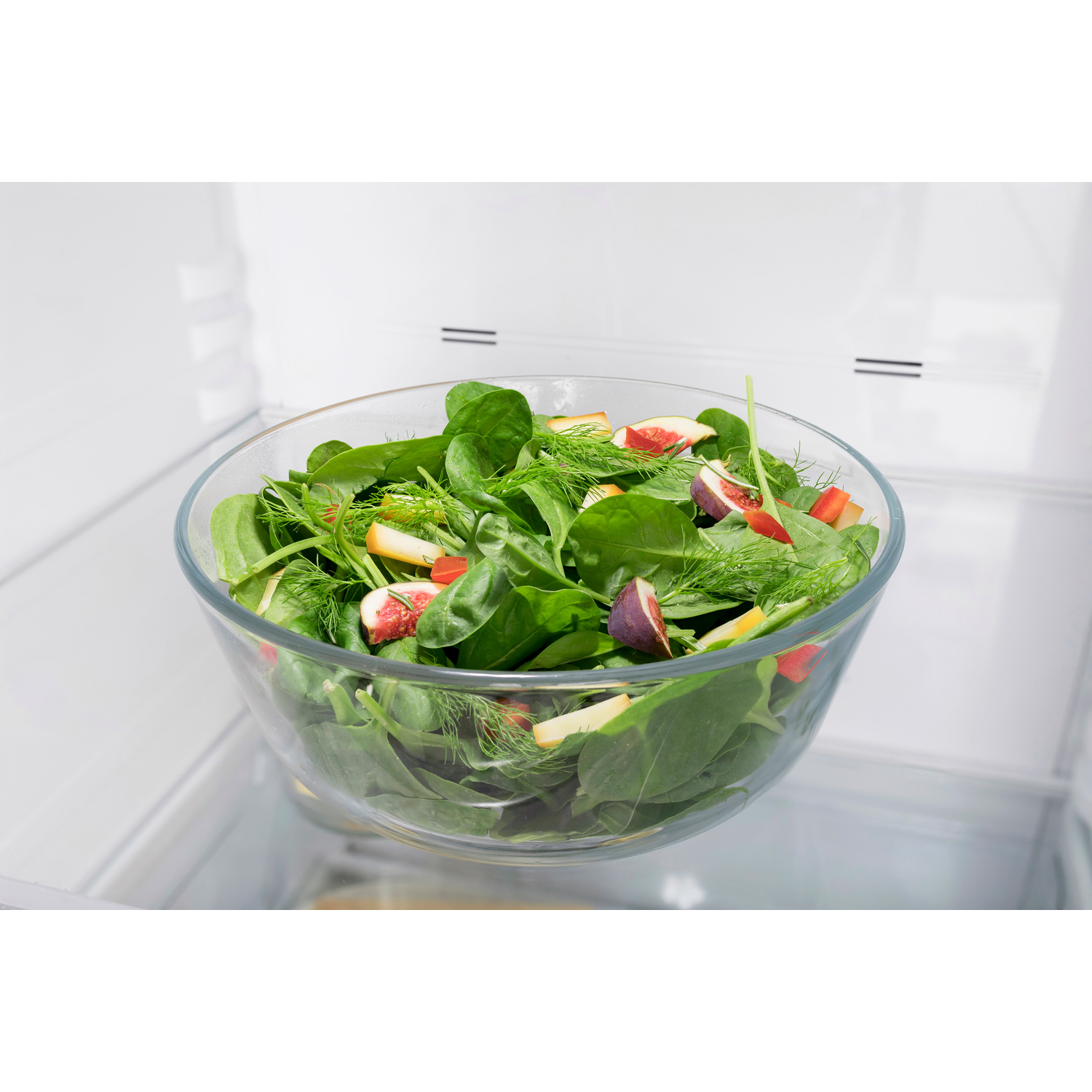 Een glazen kom met salade van spinazie, vijgen en dille staat in een koelkast.