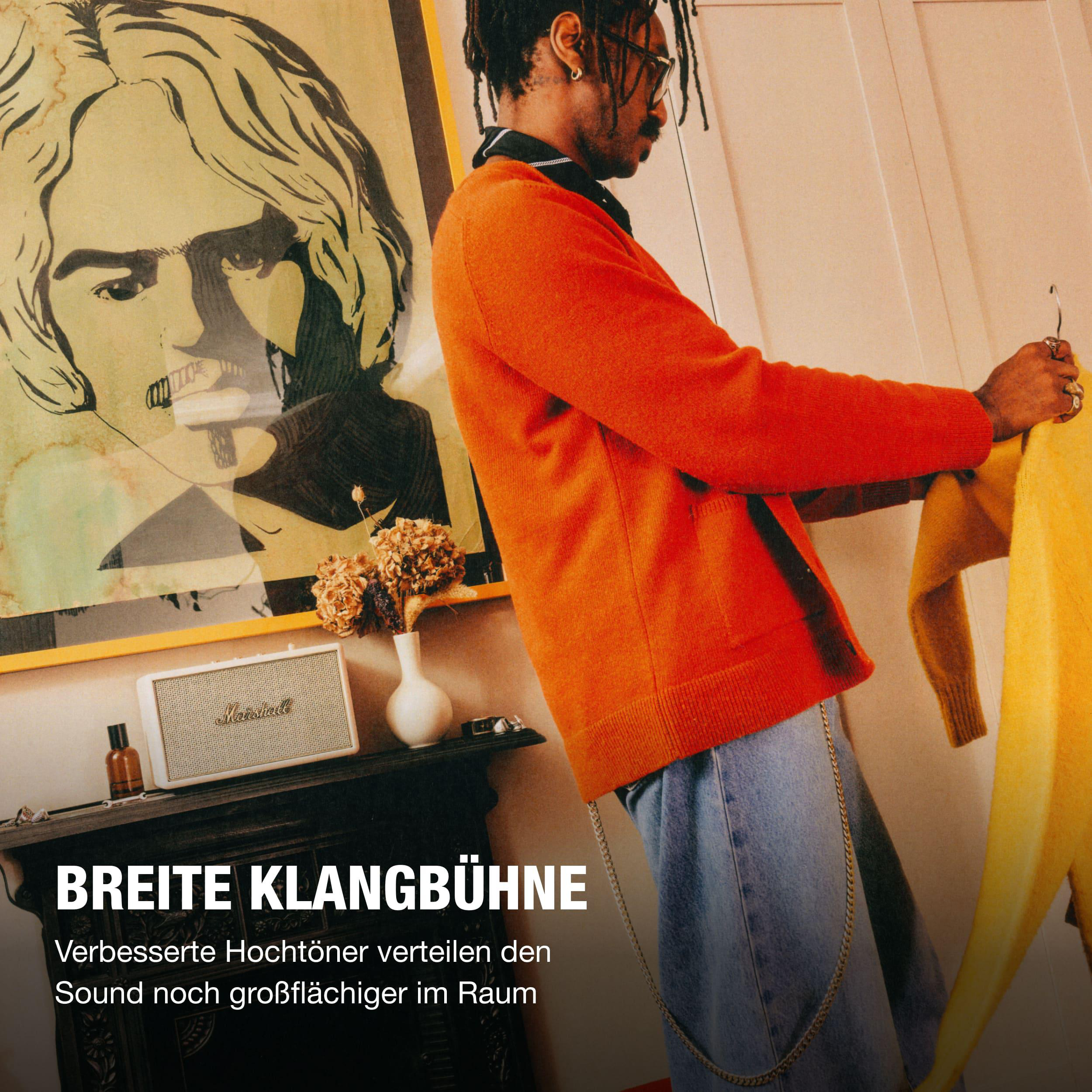Ein Mann in einem orangen Pullover steht vor einem Kunstwerk und hält ein gelbes Kleidungsstück.