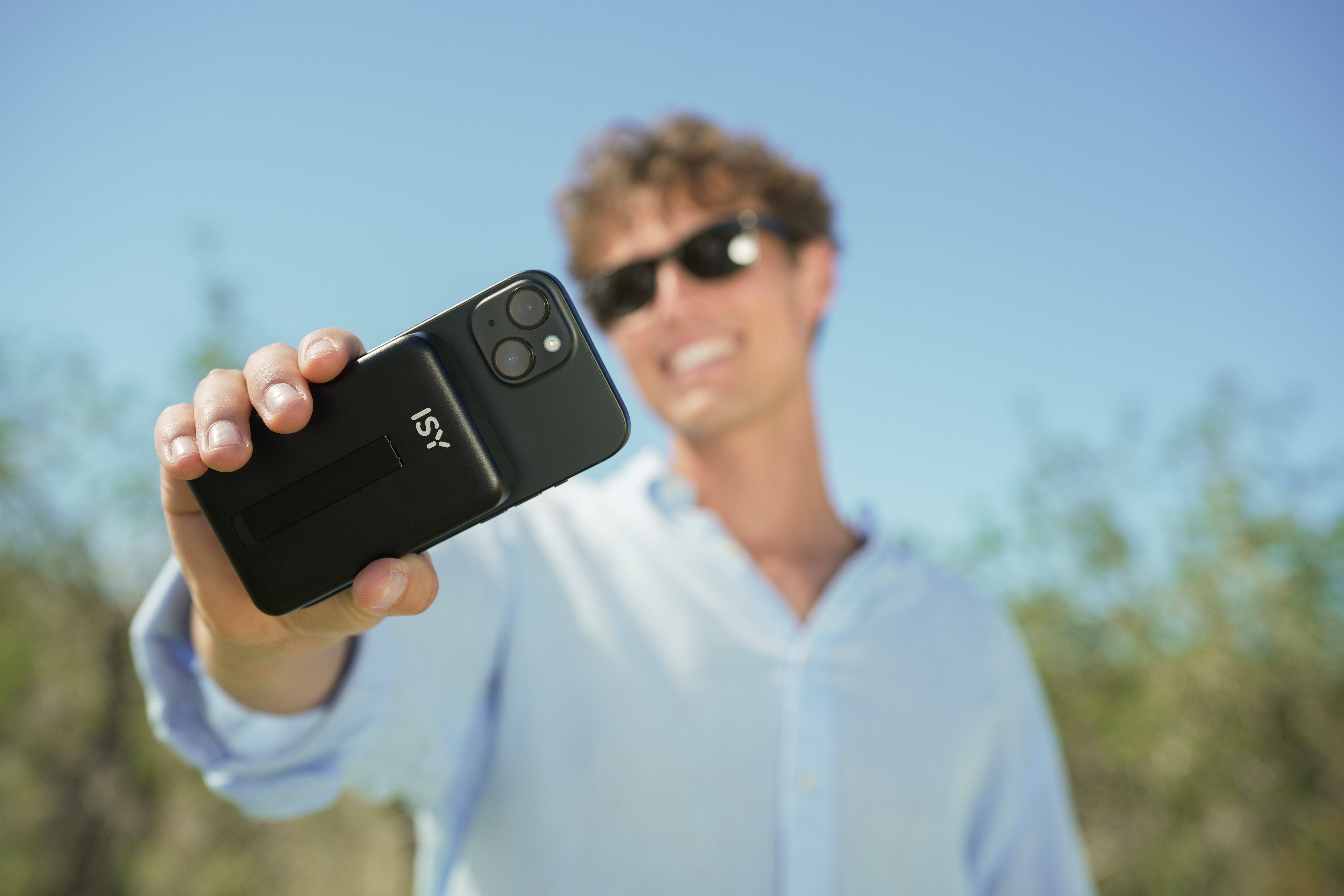 Mann hält Smartphone mit Kamera vor blauem Himmel. ISY-Logo sichtbar.